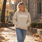 Woman wearing SIGNUMX College Hoodie in Sand, walking past a stone church in autumn. Women's Catholic collegiate hoodie lifestyle